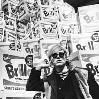 Black and white photo of a person in front of boxes of soap pads. The person is holding their hand to his or her ear.