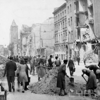 Black and white photo of destroyed buildings and civilians clearing rubble from the street; some people are just going along the street.