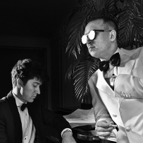 Black and white photo of two men: one sitting at the piano, another standing by it, leaning on it with his arm.