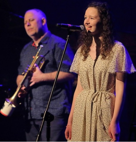Photo of the performers: young woman singing to a microphone and a man holding a trumpet in his hands.