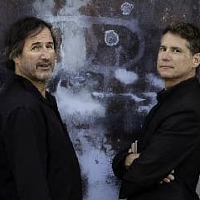 Photo of two men dressed in black clothes, who are standing opposite each other and looking to the side, into the camera.