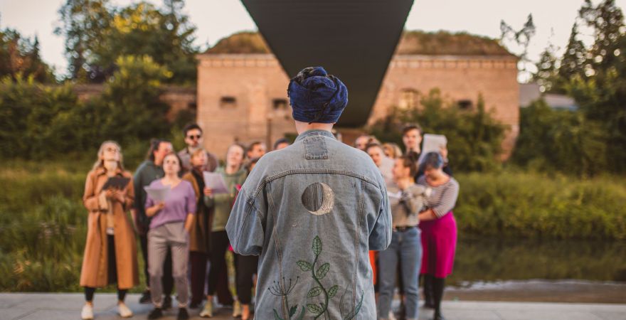 Photo of a group of singing people. In the foreground, one person with his back turned back. Green bushes and trees, and a building as a background.