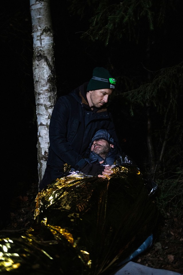 In the photo there are two men in the forest. One of them is wrapped in a gold and silver thermal emergency blanket and has her eyes closed. The other, standing above him with a headlamp on his head, holds him.