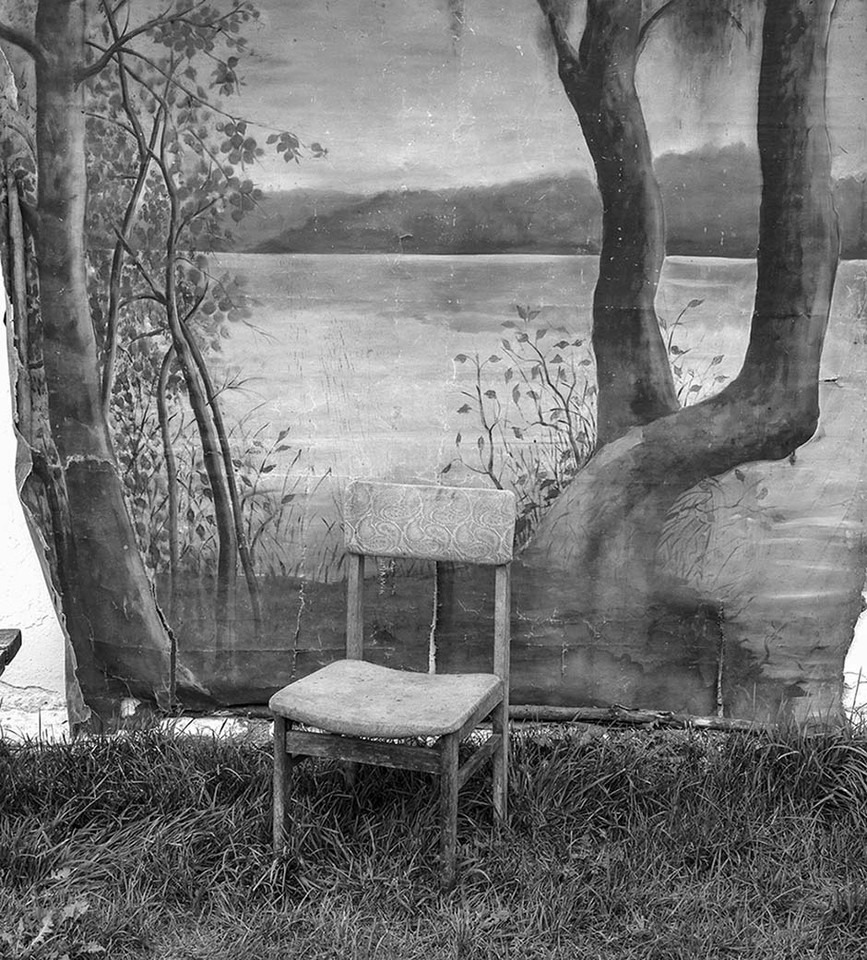 Black and white photo shows a chair standing on the ground covered with grass. Behind the chair there is an old photographic background with painted trees, water and blurry mountains on the horizon.