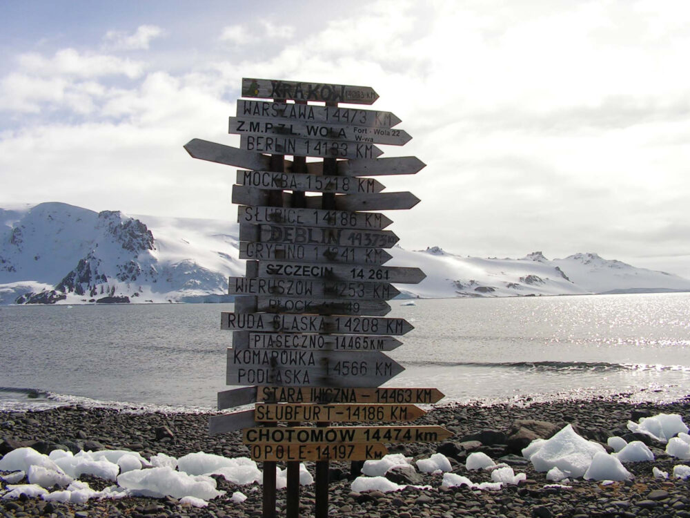 One of the exhibition works: a photo of a pole with many signposts with names of places and distances to them. In the background water and snowy mountains.