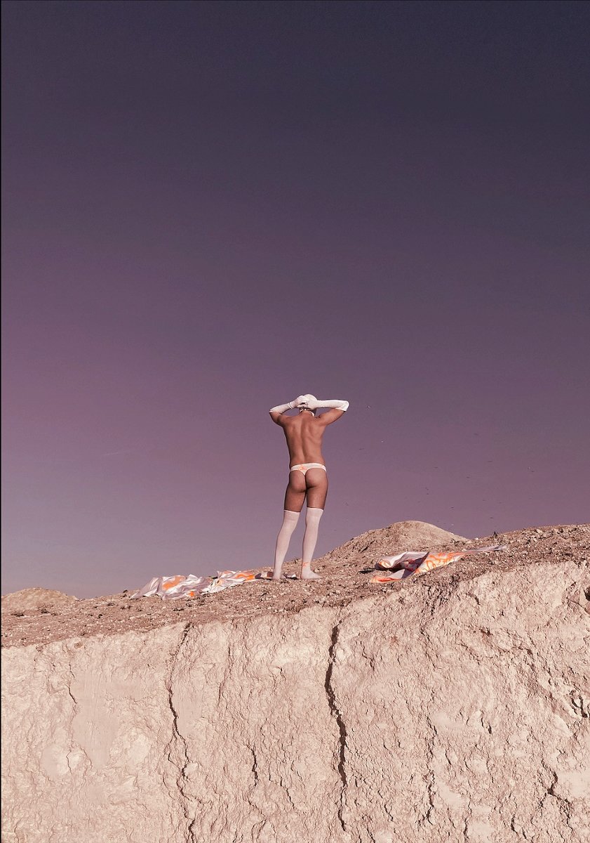 A picture of half-naked man standing on a rock, wearing white knee socks, long white gloves, white cap and white thong.