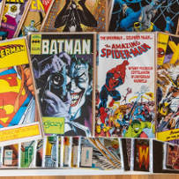 Four full color covers of Superman, Batman, Spider-Man, and X-men comics. They lie on a stack of many other comics.
