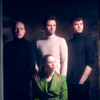 Photo of the band members - three men who are standing behind a woman who is sitting