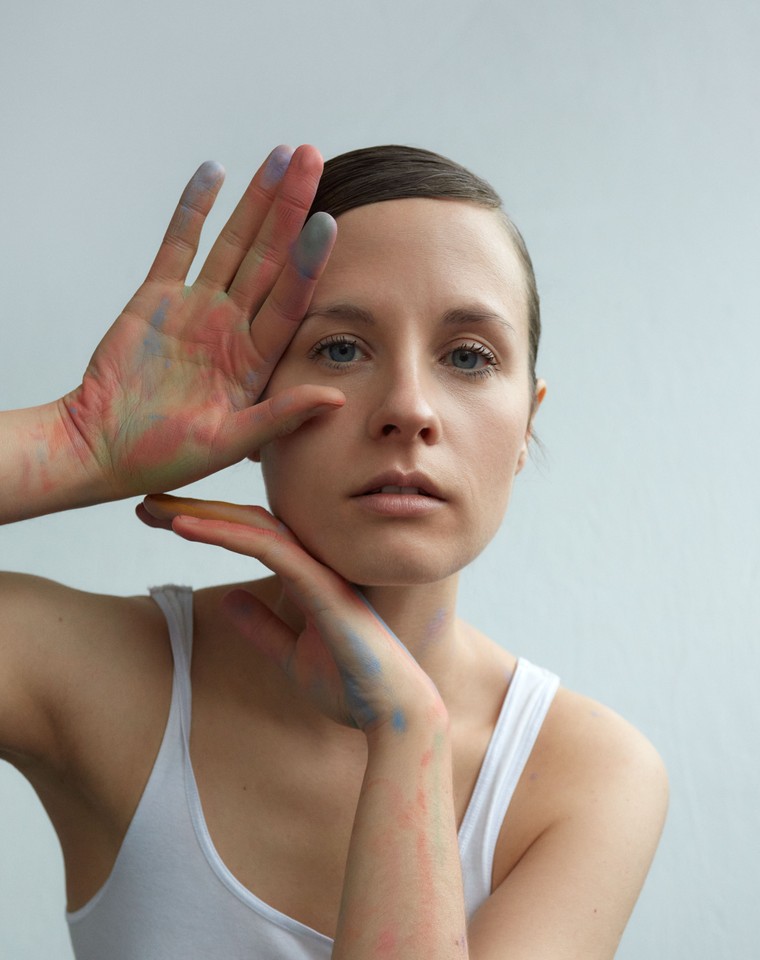 The photo shows a portrait of the artist in a white T-shirt. Her hair smoothly combed back. She touches her face with her hands slightly stained with chalk.