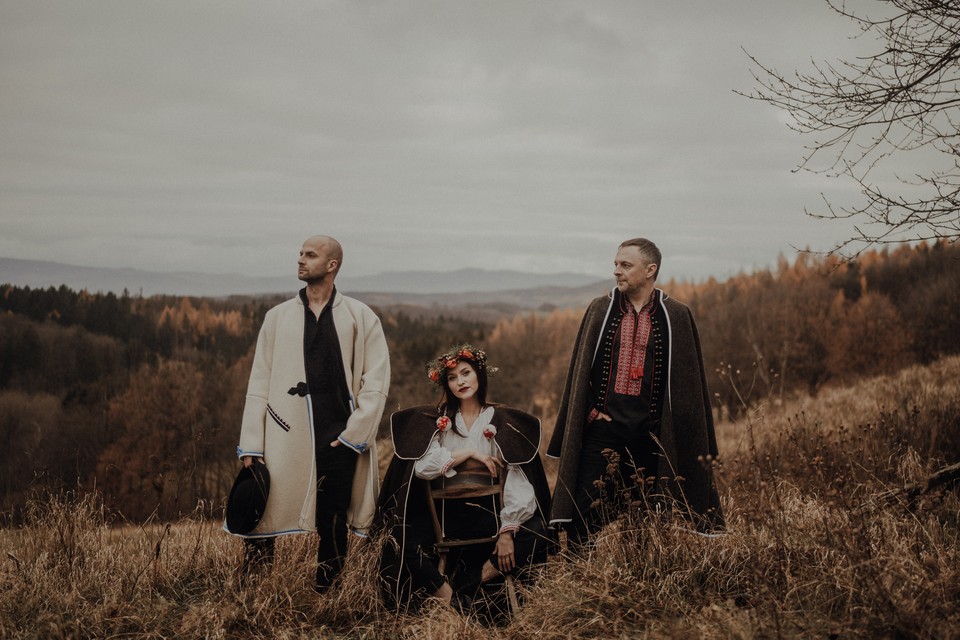 Photo of the band: a woman sitting and two men standing in folk clothes; mountainous landscape in the background