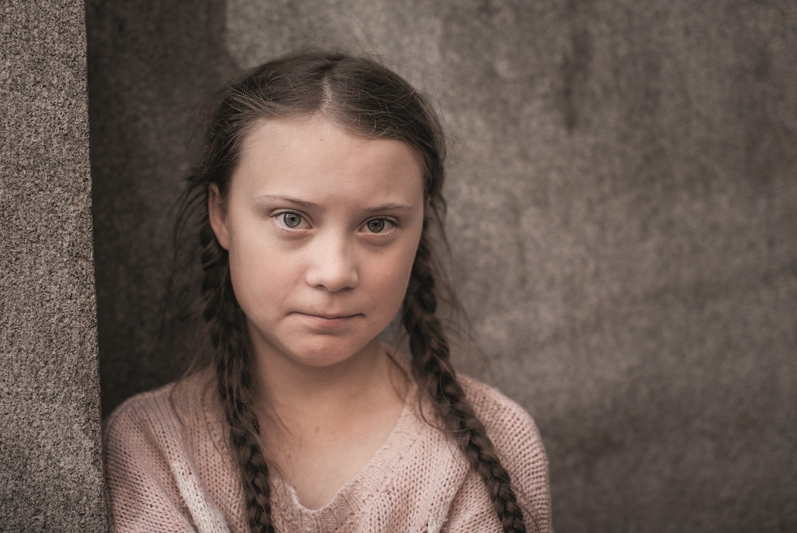 Picture of a girl in braids who is looking into the camera. Grey background.