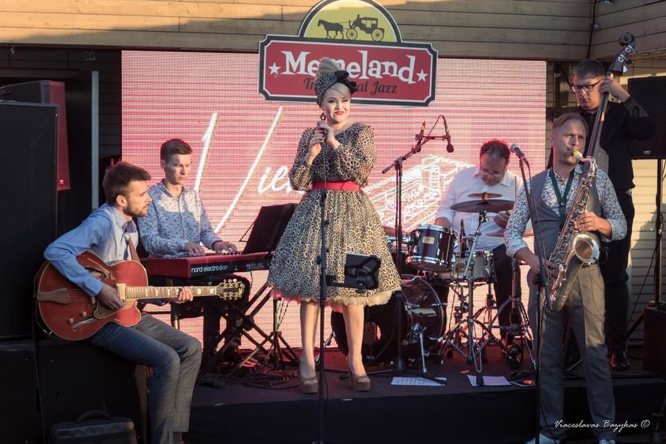 Photo of the performers on stage - a woman in a dress who is standing and holding a microphone in her hands and four men sitting and playing the musical instruments.
