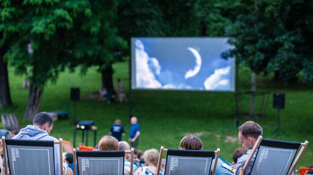 Picture presents some people sitting in a park on deck chairs, watching a film on a big screen
