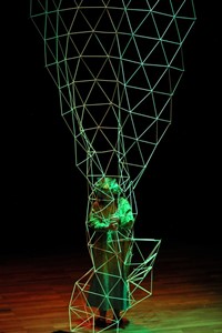 Photo presents an actor in green garment inside a green openwork wire construction on black background