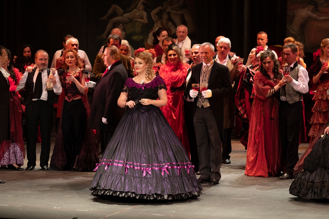 Photo from the spectacle: a woman dressed in a ball gown,many people around her, some of them with wine glasses in their hands.