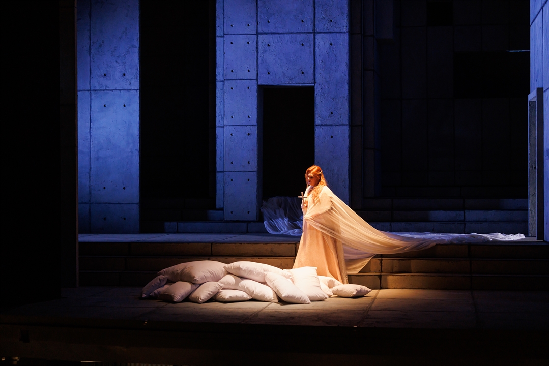 Photo from performance: a woman in a long, white dress standing on stage. She holds a cross in her hand. She stands next to several white pillows on the floor. Black and dark blue background.
