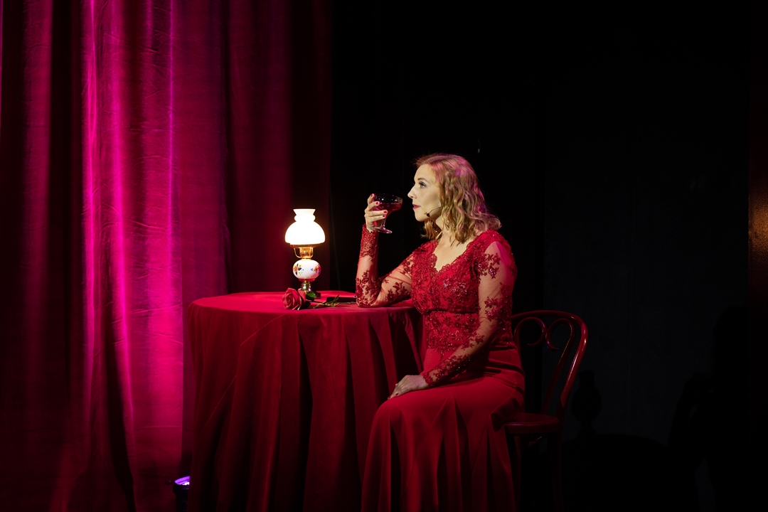 Photo of a woman in red dress, sitting at the table with a glass of champagne. The table is covered by dark red table-cloth, red curtains as a background.