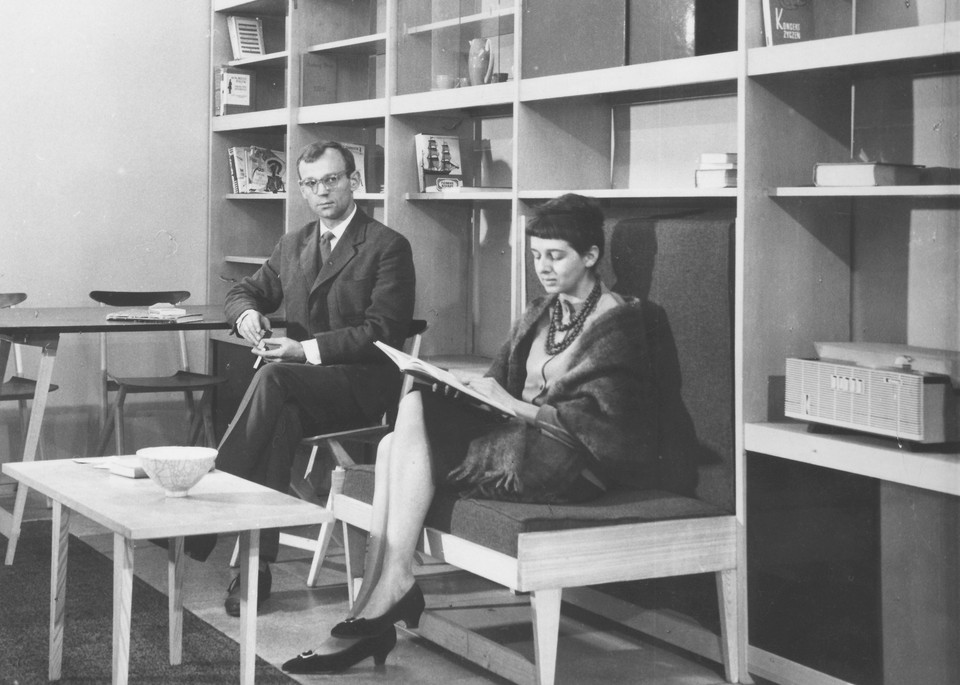 Black and white photo - a woman sitting and reading a book and a man sitting by a table, looking into the camera. In the foreground a small, low table with a ceramic bowl on it. In the background shelves with single books on them.