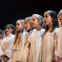 Photo of a few-year-old girls in white dresses, standing in a row and singing.