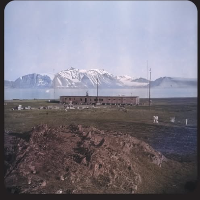 Photo of a single-story, elongated building, with water and a mountain partially covered with snow in the background. Information about the exhibition at the bottom of the photo.