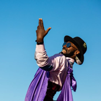 Photo of a smiling Afro-American, in sun-glasses and a black hat, The man dressed in white shirt and a violet shawl is standing sideways with his one hand raised. Blue sky as a background.
