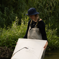 Photo of a woman, who stands in a river and holds a board with a sheet of paper. Oposite her, there is a construction consisting of a white box and a kind of metalic pointer touching the board with a paper.