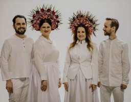 The photo shows four smiling musicians from the Dagadana band. Everyone in stylized white costume, with colorful wreaths of flowers on their heads, white wall as a background