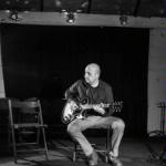 Black and white photo of a man sitting on chair and playing the guitar