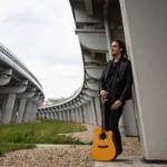 A man leaning against bridge pillar, holding a guitar