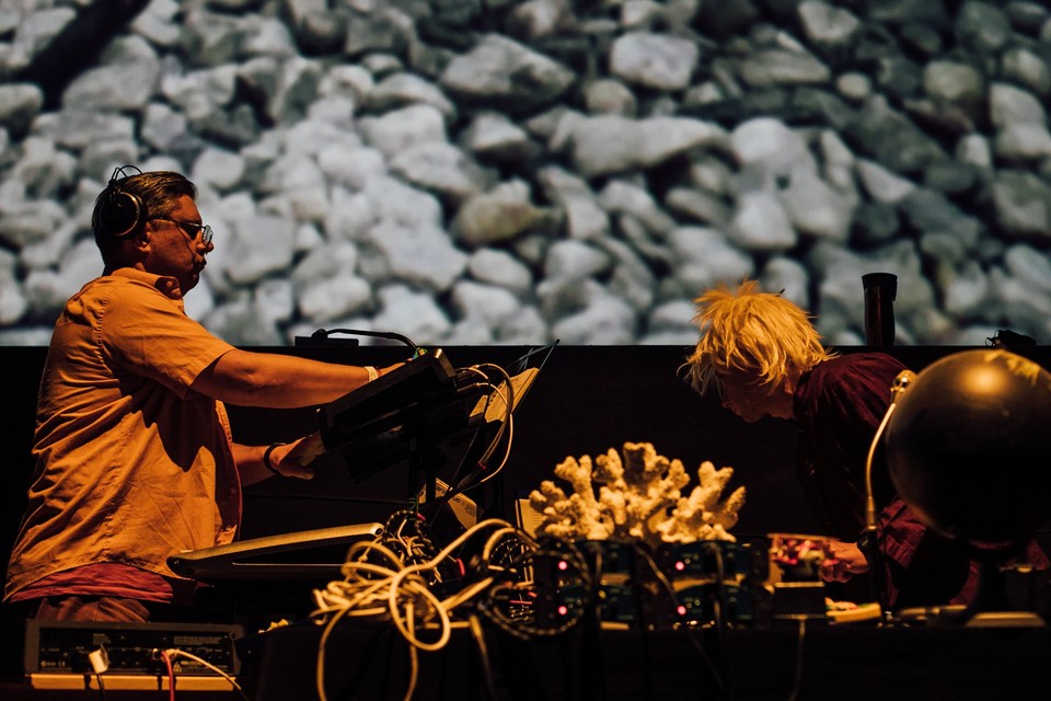 The photo shows the musicians during a performance, leaning over electronic instruments