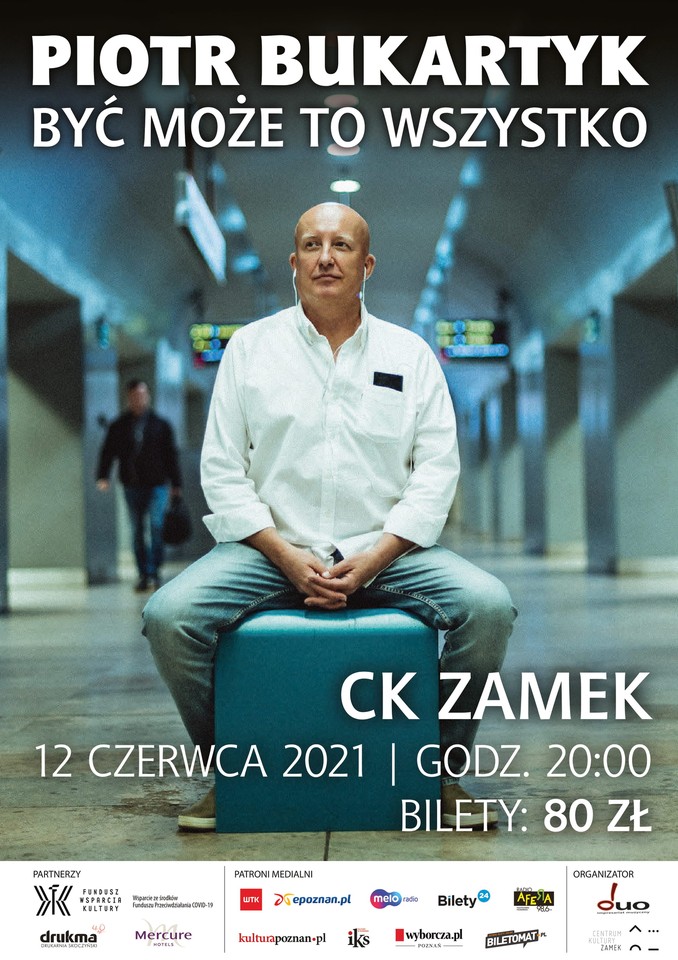 Photo of the artist in a white shirt and headphones, who is sitting on a turquoise pouf in a public space, probably in the underground passage of a train station or subway