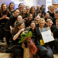 Photo of the choir members, young women and men, standing or in a squat, smiling. One of the women is holding bunch of flowers, the other is holding a diploma.