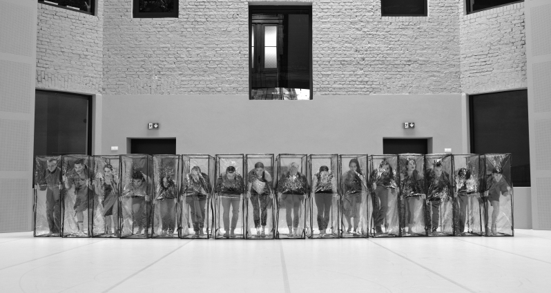 Black and white photo of the dancers in a row, who are standing in transparent boxes.