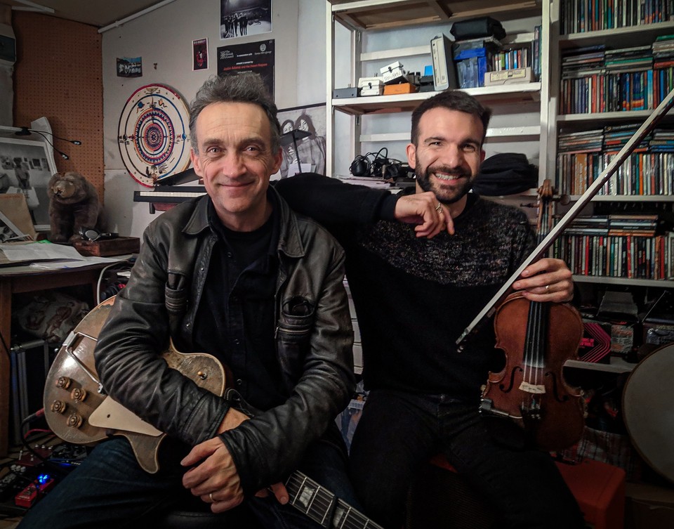 Photo of the artists: two men looking into the camera, smiling and holding their musical instruments. Shelves with CDs and various things in the background