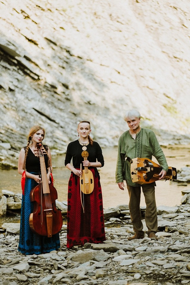 The photo shows three musicians, two women and a man, in the background there is a rocky hill. Musicians stand in front of the camera holding musical instruments