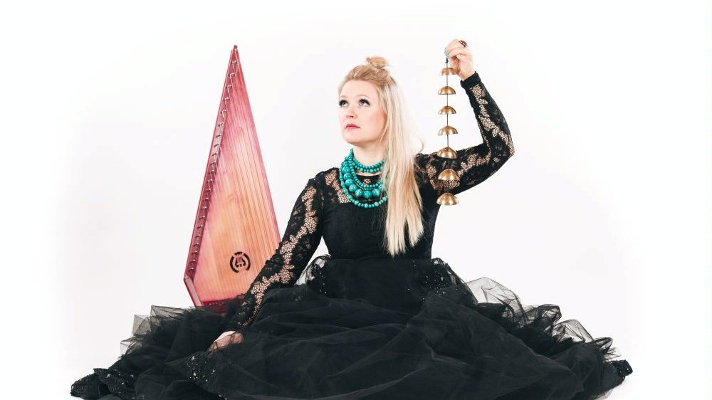 Photograph shows the artist. The woman is wearing a black dress. The sleeves are made of lace and the bottom is made of tulle. Light straight hair falls to the left shoulder. The artist raises her head slightly and looks beyond the frame. She is accompanied by musical instruments. White background.