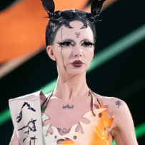 Portrait of a non-binary person with black stretchy pigtails. She is wearing a paper bra and a sash. She has a rainbow pin attached to her bra.