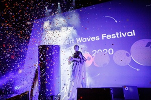 A woman standing on a stage. A big screen in the background. The picture in blue and violet colours, in front of the stage spread colourful confetti.