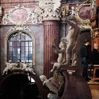 The interior of the parish church. At the top, by the side organs, sits a musician, behind him stands his companion. On the balustrades stand beautiful figures of cherubs, on the walls around hang ornate paintings of the "Stations of the Cross".