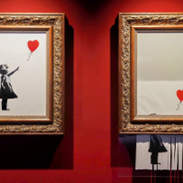 The same picture of a little girl releasing a heart-shaped balloon, placed in gold frames hanging on a red wall. On the left the picture is intact, on the right it is partially destroyed by a paper shredder.