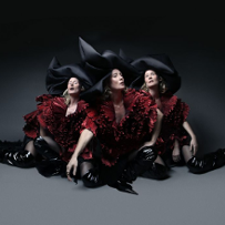 Three women are sitting on the floor. They are wearing burgundy dresses, black high boots, and black hats. Dark gray background.