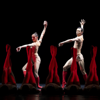 Two dancers illuminated by the light on the stage, in front of them dancers lying on the floor with their straight legs raised up in red tights.