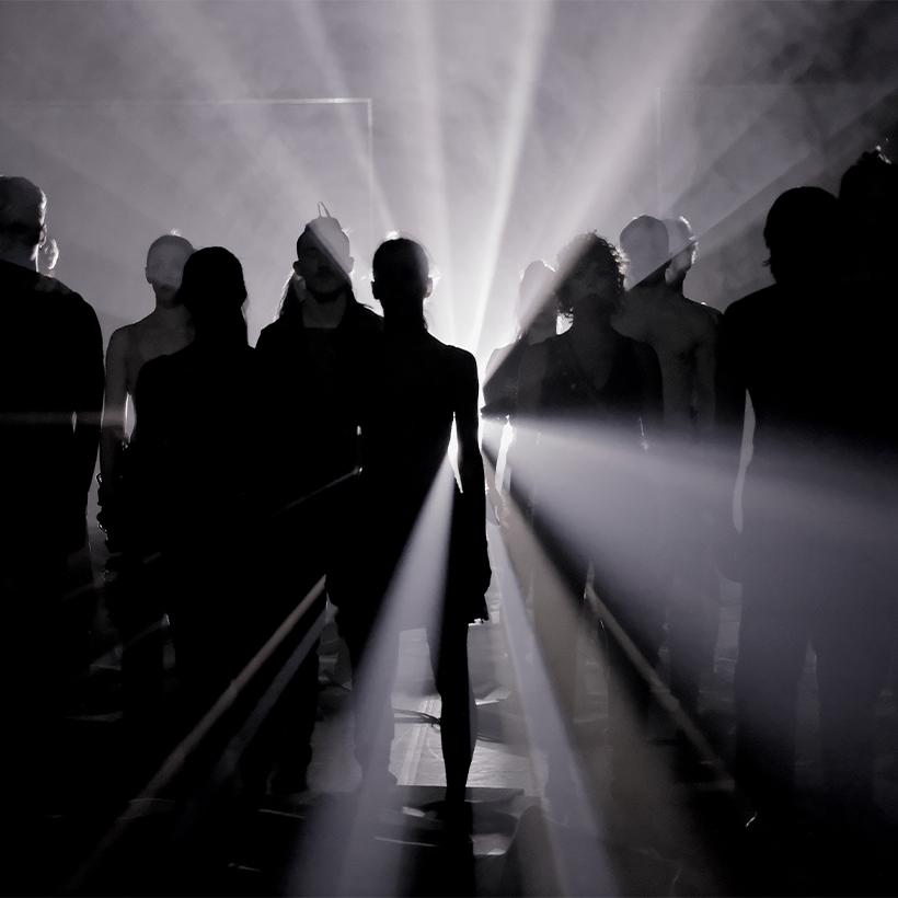 W ciemności stoi grupa taneczna, która jest oświetlona od tyłu bialym światłem.