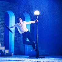 Aktor odtwarza na scenie ikoniczną scenę z Deszczowej Piosenki, tańczy stojąć na postumencie latarni ulicznej