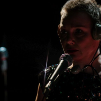 Aktorka w słuchawkach na głowie stoi przy mikrofonie w ciemności.
