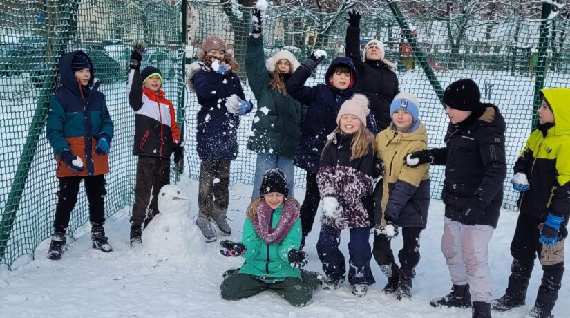 Grupa roześmianych dzieci pozuje do zdjęcia. Dzieci są ubrane w zimowe kurtki, czapki i rękawiczki. Bawią się śniegiem, podskakują i lepią bałwana.