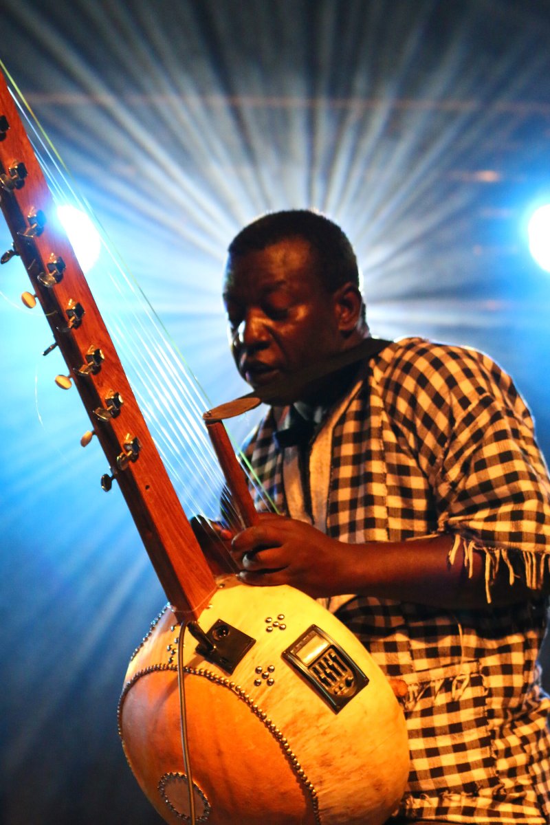 Mężczyzna w koszuli w kratę grający na korze - instrument przypomina gitarę. Za nim światła sceniczne.