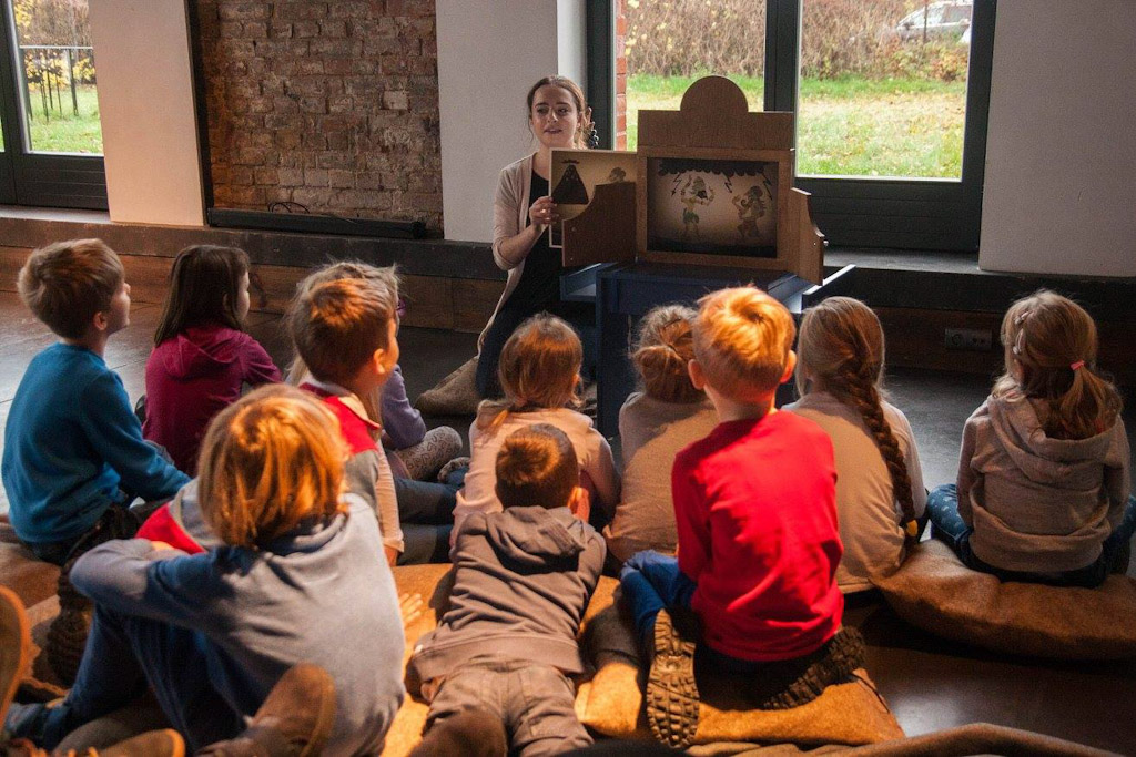 Zdjęcie, aktorka klęczy przed teatrzykiem postawionym na stole, przed nią siedzą i leżą dzieci.