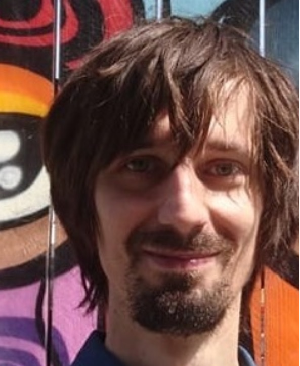 Fotografia młodego mężczyzny, Tomasza S. Markiewki, na tle kolorowego muralu w fantazyjne kształty. Markiewka ma dłuższe, brązowe włosy, zarost, uśmiecha się. Ubrany jest w niebieską, dżinsową koszulę, na plecach ma plecak.