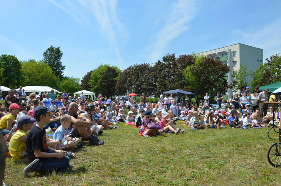 Grupa dzieci i dorosłych siedzących na trawie. W tle drzewa, niebo i blok.